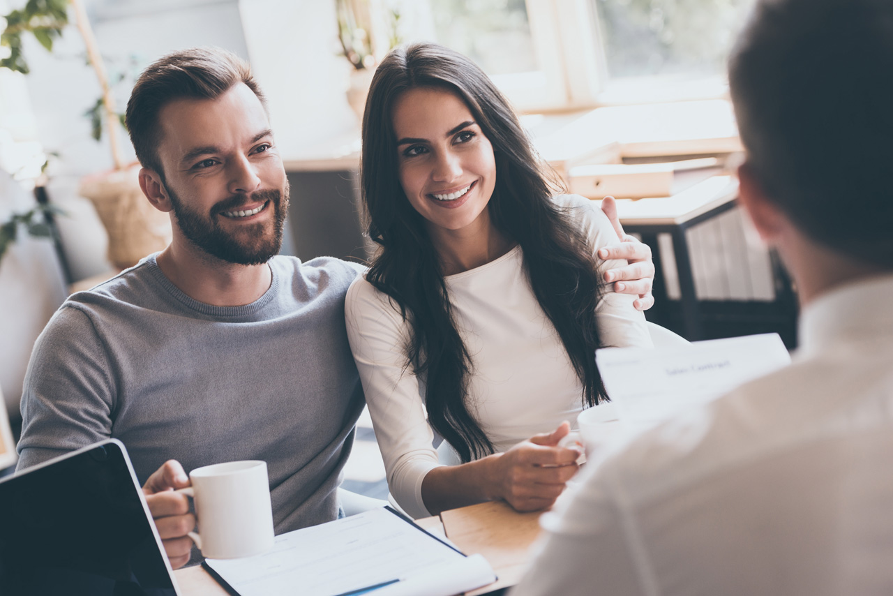 Buying something special. Young loving couple bonding to each other and looking at man sitting in front of them at the desk and holding some document